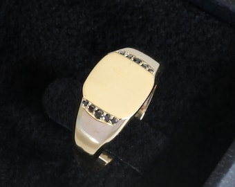 May include: A gold signet ring with a flat, rectangular top and a black stone border.