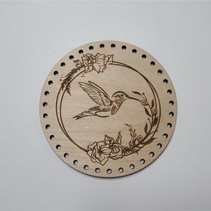 May include: A round wooden disc with a laser-etched design of a hummingbird in flight, surrounded by flowers and leaves. The disc has holes around the edge.