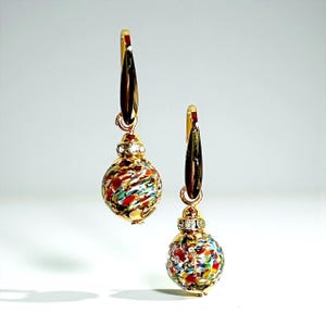 May include: A pair of gold-toned drop earrings. Each earring features a colorful, round glass bead with red, blue, and green accents, and a small, clear gem. The earrings have a decorative gold hook.