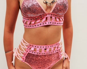 Könnte beinhalten: Zweigeteiltes Set aus rosa Pailletten mit einem Halstuch-Oberteil und einem hoch taillierten Unterteil. Beide Teile haben ein Cut-Out-Design mit silbernen Ösen und rosa Schleifenbändern.