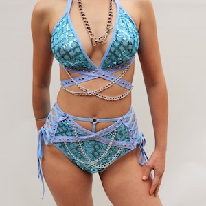 Puede incluir: Un conjunto de dos piezas azul claro con cadenas y tachuelas plateadas. La parte superior es de estilo halter con un dise&ntilde;o cruzado. La parte inferior es de cintura alta con un detalle de cordones en los lados.