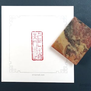 Puede incluir: Un sello rojo con caracteres coreanos sobre un fondo blanco. El sello está al lado de una pequeña piedra rectangular multicolor.