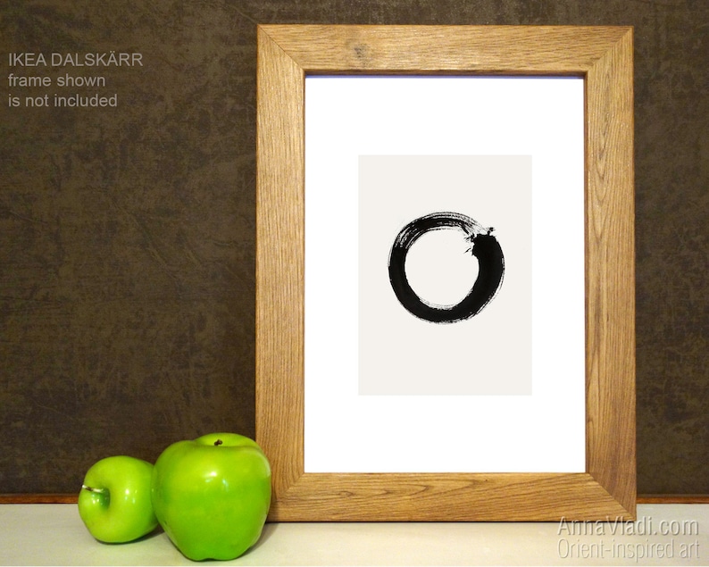 May include: A black and white Zen circle print in a wooden frame. The print is on a white background and the circle is made with black ink. The frame is a light brown wood.