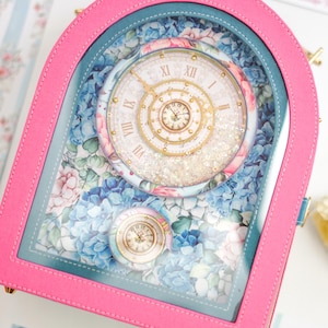 May include: A pink and teal decorative clock with a clear front, featuring a floral design and a clock face with Roman numerals. The clock has a small, round clock face at the bottom and is surrounded by a floral pattern.