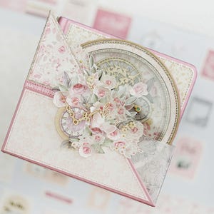 Pode incluir: Uma caixa decorativa aberta com um design floral e de relógio. A caixa é principalmente rosa e branca, com um padrão floral e um mostrador de relógio no centro. Decorada com flores de papel e um design de bússola.