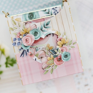 May include: A decorative box with a heart-shaped cutout, adorned with floral embellishments in shades of pink, blue, and yellow. The box features a pink striped pattern and a wooden plank design. Gold accents are visible.