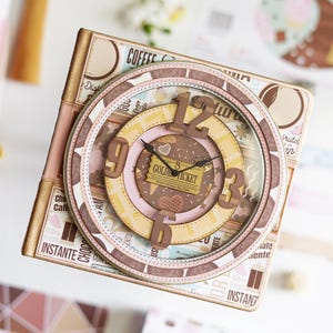May include: A decorative clock with a whimsical design. The clock face features a layered design with a brown, pink, and yellow colour scheme. The clock hands point to 10:09. The clock has the words "Golden Ticket" on the face.