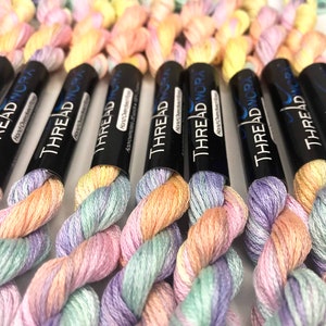 May include: Close-up of several spools of hand-dyed embroidery floss. The floss is a gradient of pastel pink, green, and yellow colors. Each spool is labeled "THREAD NORA" with the text "Hand Overdyed Floss".