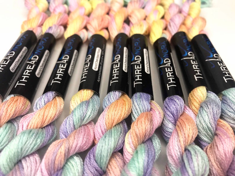 May include: Close-up of several spools of hand-dyed embroidery floss. The floss is a gradient of pastel pink, green, and yellow colors. Each spool is labeled "THREAD NORA" with the text "Hand Overdyed Floss".
