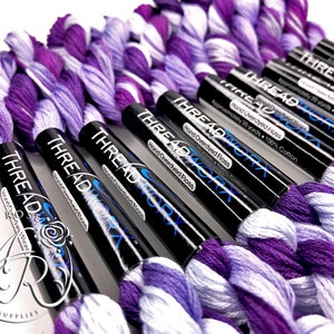 May include: A close-up of several spools of purple and white hand-dyed embroidery floss. The spools are labeled "THREAD WORX" and "Hand Overdyed Floss".