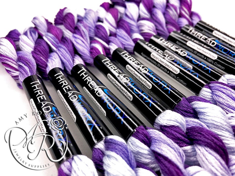 May include: A close-up of several spools of purple and white hand-dyed embroidery floss. The spools are labeled "THREAD WORX" and "Hand Overdyed Floss".
