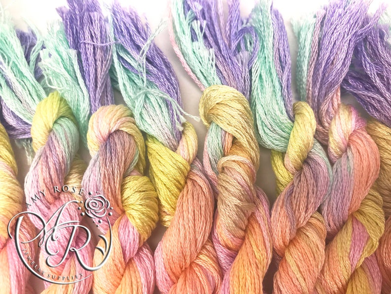 May include: Close-up of a bundle of six skeins of embroidery floss in pastel shades of purple, green, yellow, pink, and orange. The floss is twisted together and has a soft, fuzzy texture.