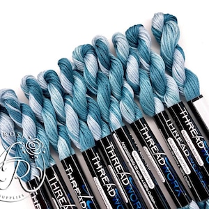May include: Close-up of several skeins of hand-dyed embroidery floss in shades of teal and white. The floss is labeled "THREAD WORK" and "1021". Each skein contains approximately 20 yards of 100% cotton thread. The image is on a white background.