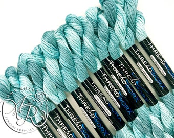 May include: A collection of teal and blue hand-dyed embroidery floss skeins. Each skein is labelled "THREAD WORX" and contains approximately 18.3 metres of 100% cotton. The image is set against a white backdrop.