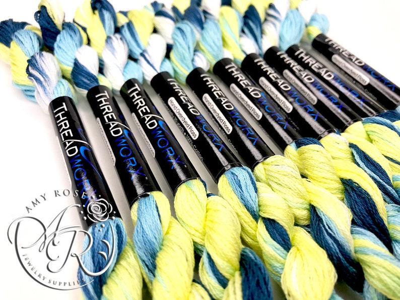 K&ouml;nnte beinhalten: Nahaufnahme von acht Spulen handgef&auml;rbter Stickgarn in Blaut&ouml;nen, Gr&uuml;n- und Gelbt&ouml;nen. Die Spulen sind mit "THREAD WORK" und "Hand Overdyed Floss" beschriftet.