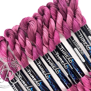 May include: A close-up of eight spools of hand-dyed embroidery floss in shades of pink and purple. The spools are labeled "THREADWORX" and "Hand Overdyed Floss".