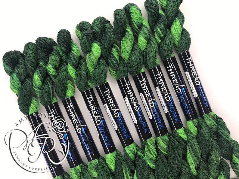 Op de afbeelding: Acht spoelen handgeverfde garen in groen en limoengroen, elke spoel met het label "THREADWORX Hand Overdyed Floss".