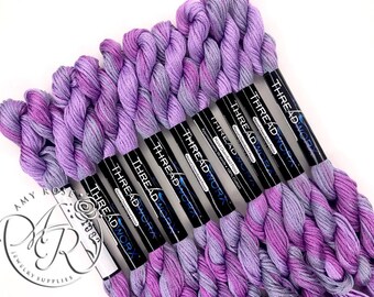 Può includere: Primo piano di otto rocchetti di filo da ricamo viola e grigio. I rocchetti sono etichettati con "THREAD WORX" e "Hand Overdyed Floss".
