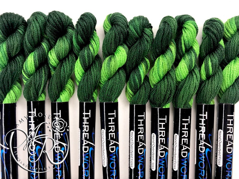 Op de afbeelding: Tien spoelen handgeverfde floss in groen en limoengroen, elk met het label "THREADWOR Hand Overdyed Floss".