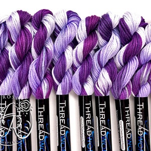 May include: A close-up of ten spools of hand-overdyed embroidery floss in shades of purple and white. The spools are labeled "THREADWOR" and "Hand Overdyed Floss".