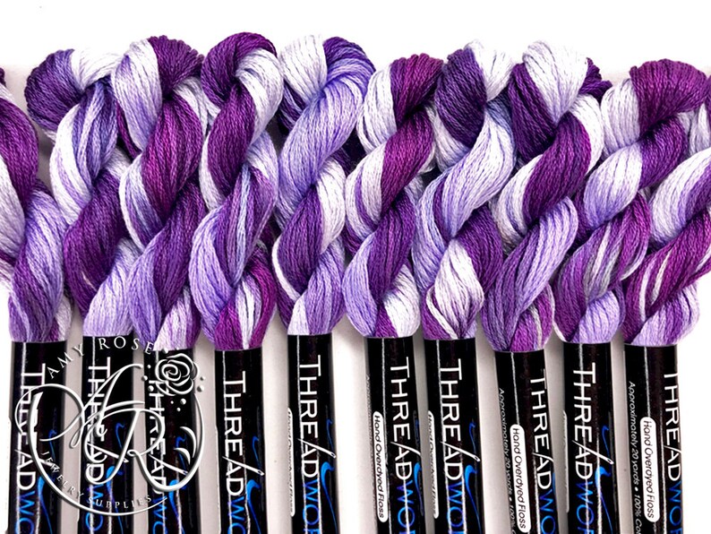 May include: A close-up of ten spools of hand-overdyed embroidery floss in shades of purple and white. The spools are labeled "THREADWOR" and "Hand Overdyed Floss".