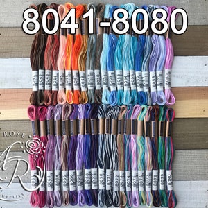 May include: A collection of embroidery floss in various colors, arranged in neat rows. The floss is labeled with numbers ranging from 8041 to 8080. The color palette includes shades of brown, orange, blue, purple, and pink. The background is a wooden surface.