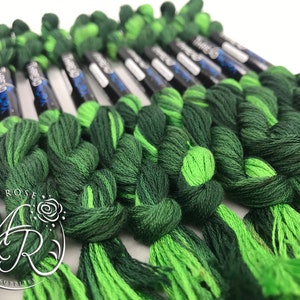 Op de afbeelding: Een close-up van een bundel groene en limoengroene borduurgaren spoelen. De spoelen zijn in een rij gerangschikt en zijn met een knoop aan elkaar gebonden. De spoelen zijn gelabeld met de tekst "DMC" en een nummer.