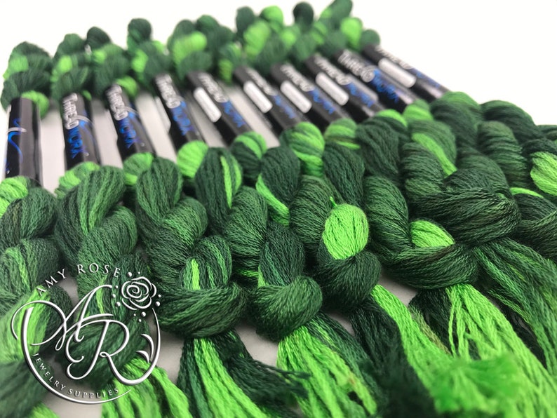 Op de afbeelding: Een close-up van een bundel groene en limoengroene borduurgaren spoelen. De spoelen zijn in een rij gerangschikt en zijn met een knoop aan elkaar gebonden. De spoelen zijn gelabeld met de tekst "DMC" en een nummer.