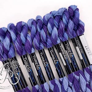 Op de afbeelding: Een verzameling handgeverfde borduurgarens in paarse en blauwe tinten. De spoelen zijn voorzien van de labels "THREAD WORX" en "10363". De garens zijn in een rij gerangschikt, waarbij de kleurvariaties worden getoond.