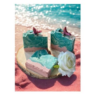 May include: Artisan soaps with a beach theme. The soaps have a teal and white swirl design, with a pink base, and are topped with a small starfish. A white rose and a wooden slice are also in the image.