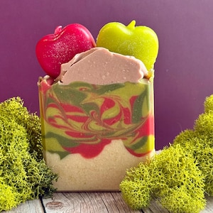 May include: A rectangular bar of soap featuring a marbled swirl of red, green, and cream colors. The top is topped with a smooth, light brown layer and two small, shiny apple-shaped soap embeds in bright red and lime green with a glittery finish.