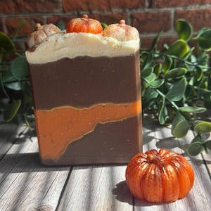 May include: A rectangular bar of soap featuring layers of dark brown and bright orange with a wavy gold line. The top is topped with white frosting-like soap and three small pumpkin-shaped soap decorations, with a separate orange pumpkin soap.