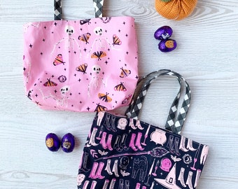 May include: Two Halloween-themed tote bags with black and white gingham handles. One pink bag features a skeleton and moth pattern, while the other is black with a witch boot design. A small orange pumpkin decoration and two foil-wrapped chocolate eggs are also visible.
