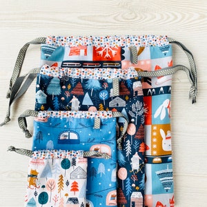 May include: Set of four drawstring bags with winter-themed fabric prints. The bags are layered on top of each other, with the top bag featuring a red, white and blue design with a sled and snowflakes. The second bag features a blue and white design with a forest scene. The third bag features a blue and white design with a cabin and trees. The fourth bag features a white and pink design with a forest scene.