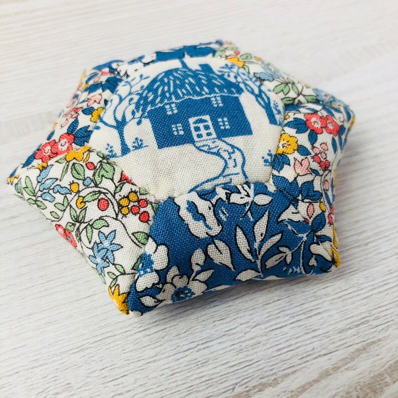 May include: A hexagonal pincushion with a blue and white floral pattern. The center panel features a white house with a blue roof and a blue door.