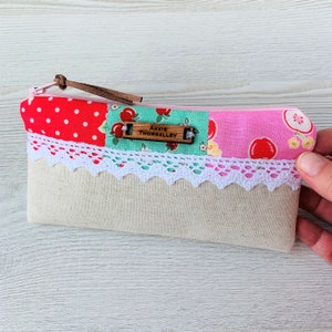 May include: A small, rectangular zippered pouch made of beige linen with a patchwork design. The patchwork includes red with white polka dots, green with white flowers and red apples, and pink with white flowers and red apples. The pouch has a white lace trim and a wooden tag with the name "Annie Thornalley" written on it.
