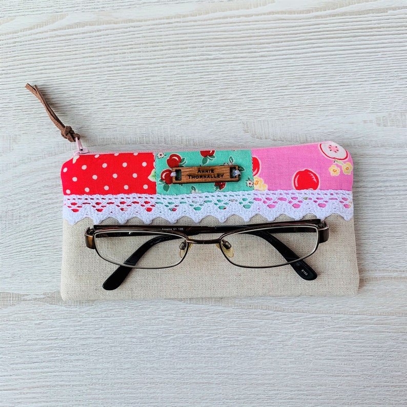 May include: A beige fabric glasses case with a patchwork design of red polka dots, green and white floral print, and pink with white polka dots. The case has a white lace trim and a wooden tag that reads "Annie Thornalley".