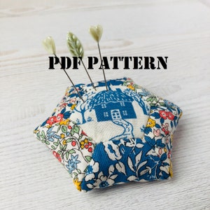 May include: A hexagonal pincushion with a blue and white floral pattern. The pincushion has a small house design in the center and three heart-shaped pins.