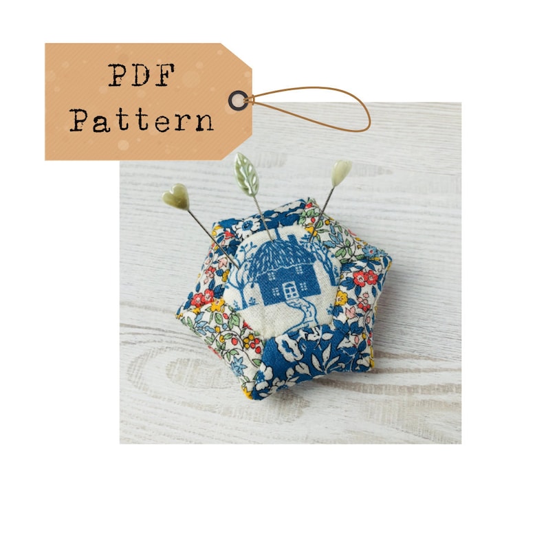 May include: A blue and white fabric pincushion shaped like a house with a thatched roof. The pincushion has a floral pattern and is decorated with green leaves and heart-shaped pins.