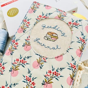 Könnte beinhalten: Ein weißer Stoff-Journal-Umschlag mit einem rosafarbenen Blumenmuster. Der Umschlag hat einen runden gestickten Aufnäher mit dem Text "Reading Journal" und einem Stapel Bücher.
