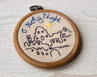 May include: A wooden embroidery hoop with a beige linen fabric inside. The fabric is embroidered with a black outline of a city skyline and a gold star. The text "O Holy Night" is embroidered in blue thread above the skyline.