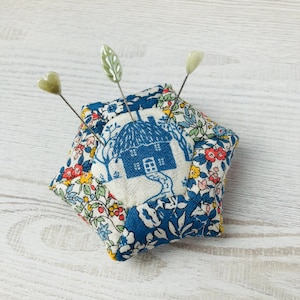 May include: A blue and white floral fabric pincushion shaped like a star with a small house design in the centre. Three decorative pins are stuck in the pincushion.