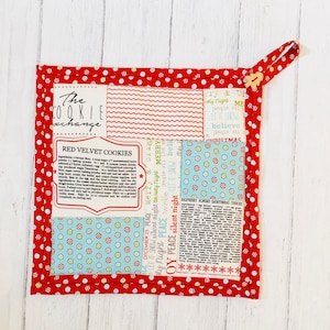 Op de afbeelding: Een gewatteerde pannenlap met een rode stippelrand. Het midden heeft stoffen panelen met tekst en patronen, waaronder een recept voor "Red Velvet Cookies" en vakantie zinnen. Een houten knoop zekert de ophanglus.