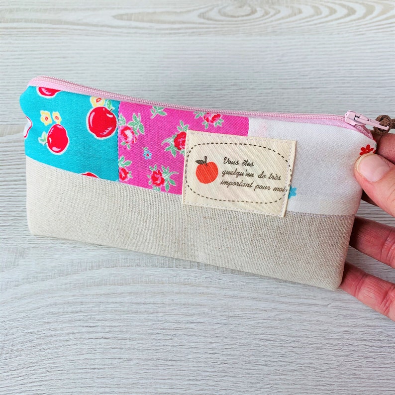 May include: A pink and blue patchwork fabric pencil case with a zipper closure. The case has a white label with a red apple and the text "Vous &ecirc;tes quelqu'un de tr&egrave;s important pour moi".