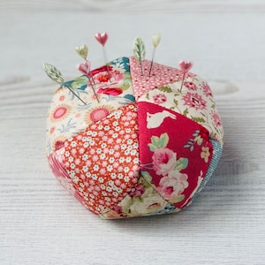 May include: A patchwork pincushion in shades of pink, red, and white with a floral pattern. The pincushion is filled with pins with heart-shaped heads.