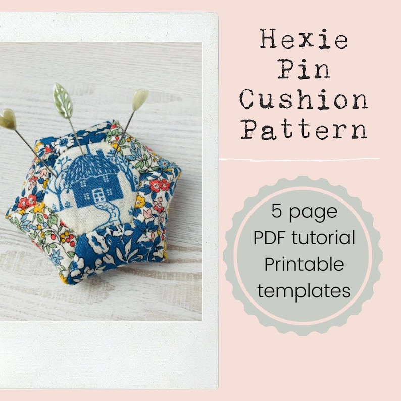 May include: A blue and white floral fabric pincushion shaped like a hexagon with a small house design in the center. The pincushion has three pins stuck in it. The pincushion is sitting on a white wooden surface. The text "Hexie Pin Cushion Pattern" and "5 page PDF tutorial Printable templates" is visible in the image.