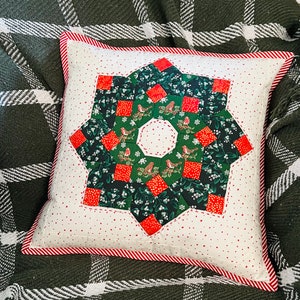 May include: Square decorative pillow with a starburst design. The center features a wreath of birds and greenery, surrounded by red squares and a white background with red polka dots. The pillow has a red and white striped border.