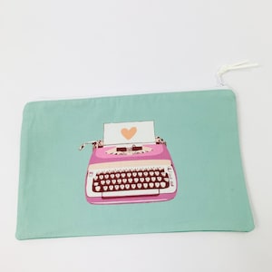 Puede incluir: Una bolsa de cremallera azul claro con una máquina de escribir rosa y un corazón en un papel blanco. La máquina de escribir tiene una palanca de retorno de carro roja y un teclado negro.