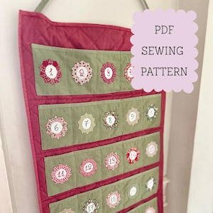Peut inclure: Calendrier de l'Avent en tissu rouge et vert, avec des poches numérotées et des ornements floraux. Le texte "PDF SEWING PATTERN" est dans un nuage violet clair. Le motif matelassé ajoute de la texture.