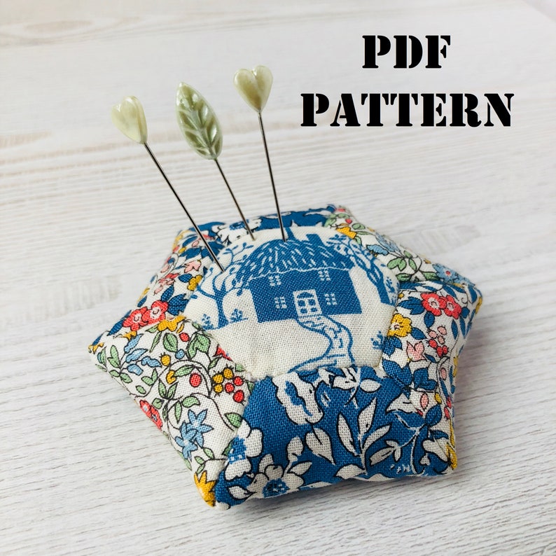 May include: A hexagonal pincushion with a blue and white floral pattern. The pincushion is filled with stuffing and has three pins sticking out of it. The pins have green and white plastic tops in the shape of leaves and a heart. The text "PDF PATTERN" is printed in black letters on a white background.